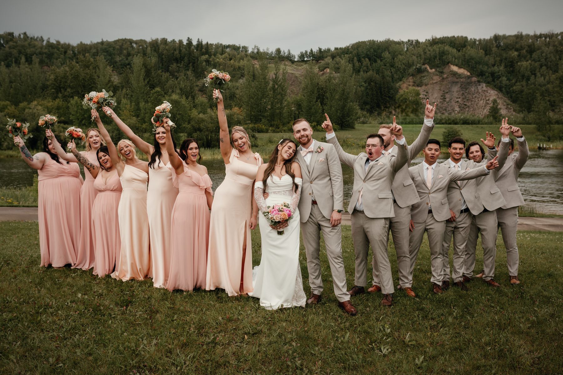 The wedding party cheering on the lawn above the lake, bouquets raised