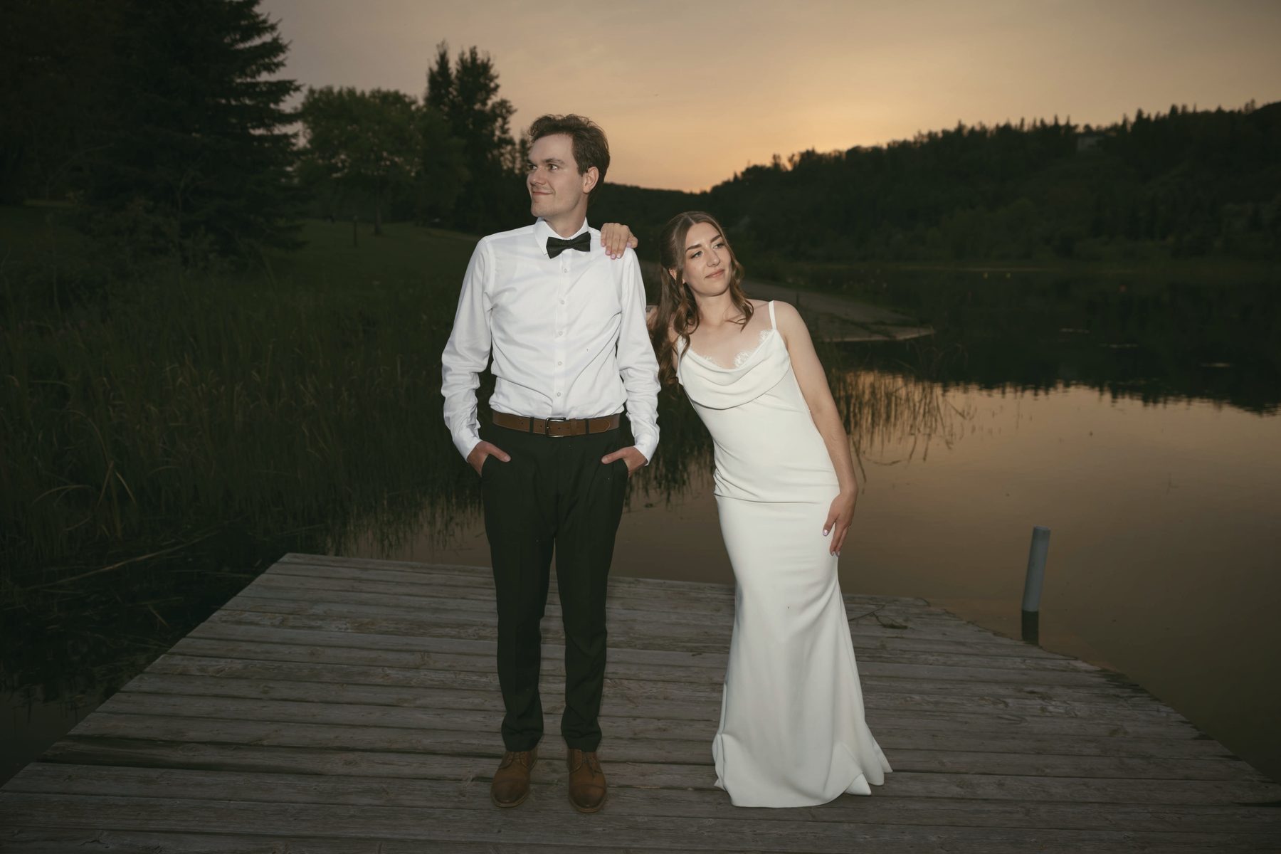 A couple standing on the dock at dusk, the water calm behind them