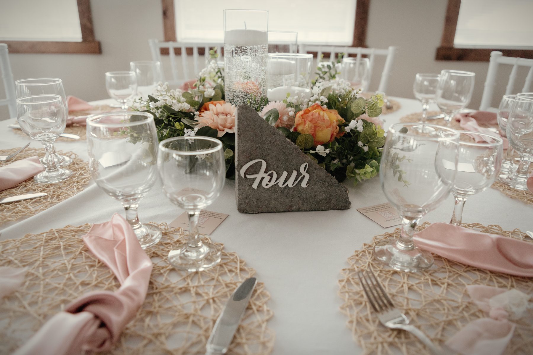 A table set with woven chargers, blush napkins, stemmed glasses and a stone table number in a nest of florals