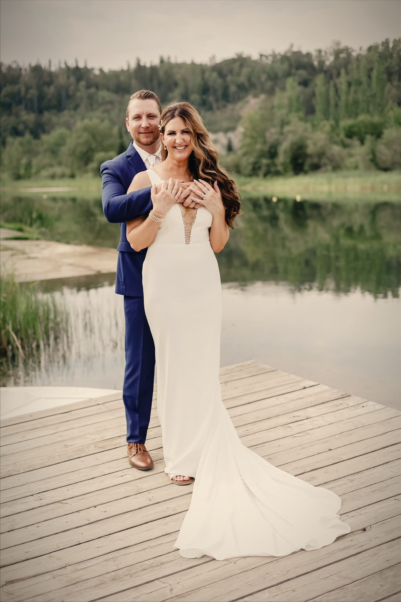 A groom holding his bride from behind on the dock, her hands over his, the lake and reeds beyond