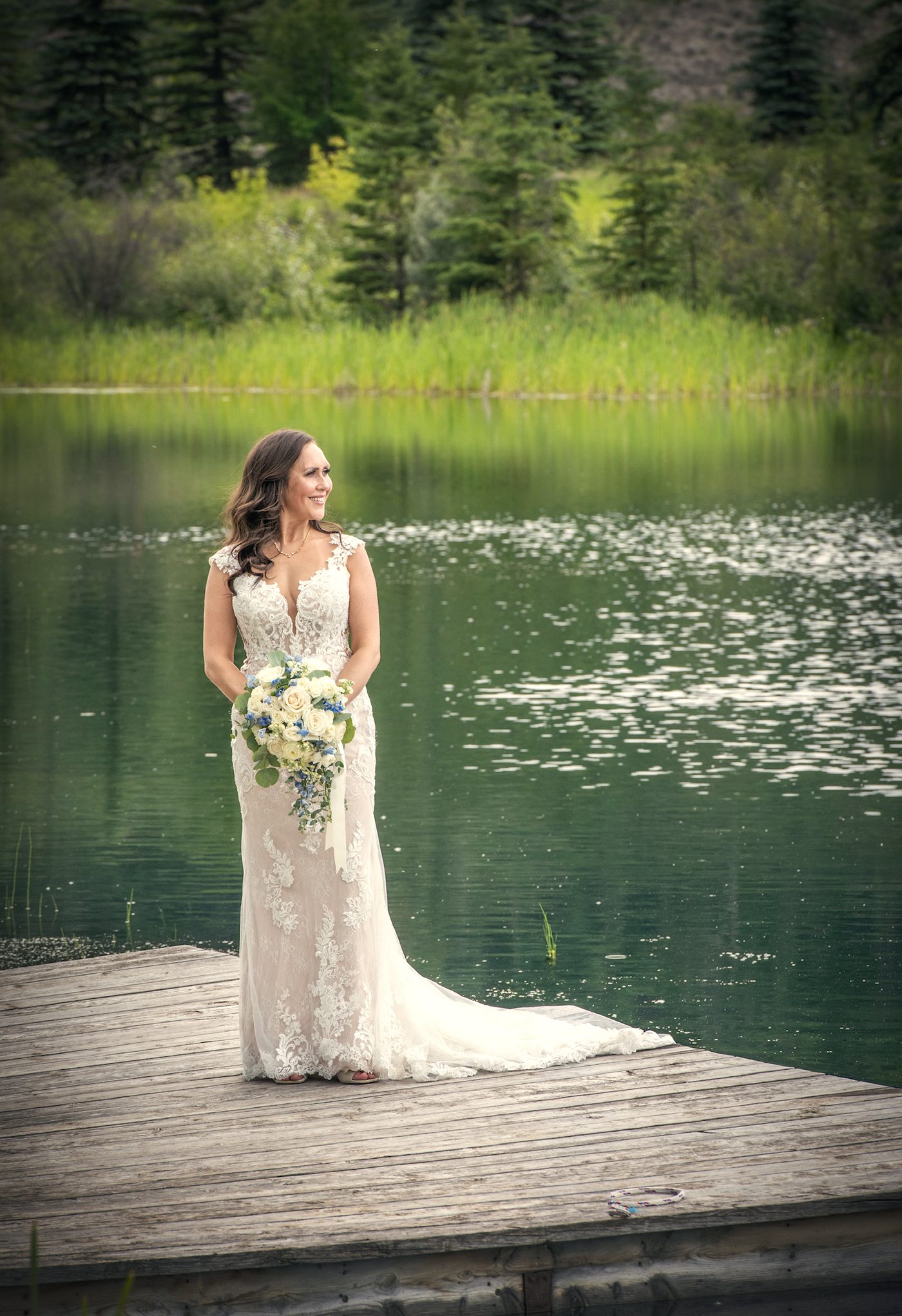 A bride alone on the dock with her bouquet, green water and floating blossom behind her