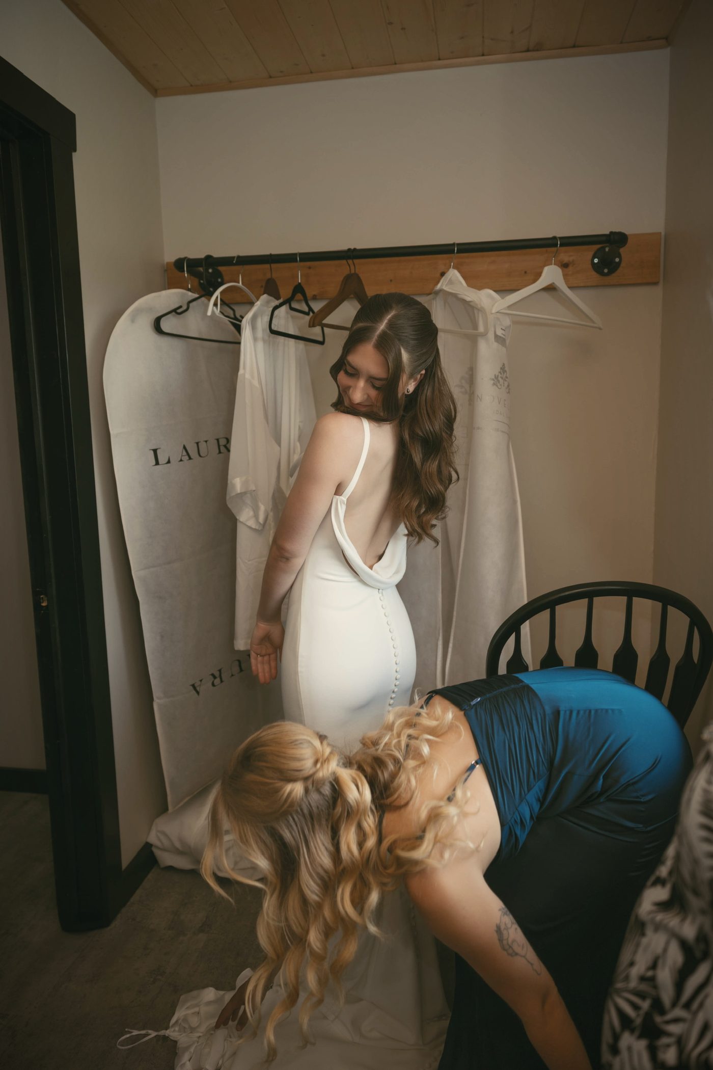 Two bridesmaids fastening a bride's dress in the suite, gowns hanging on the wall behind