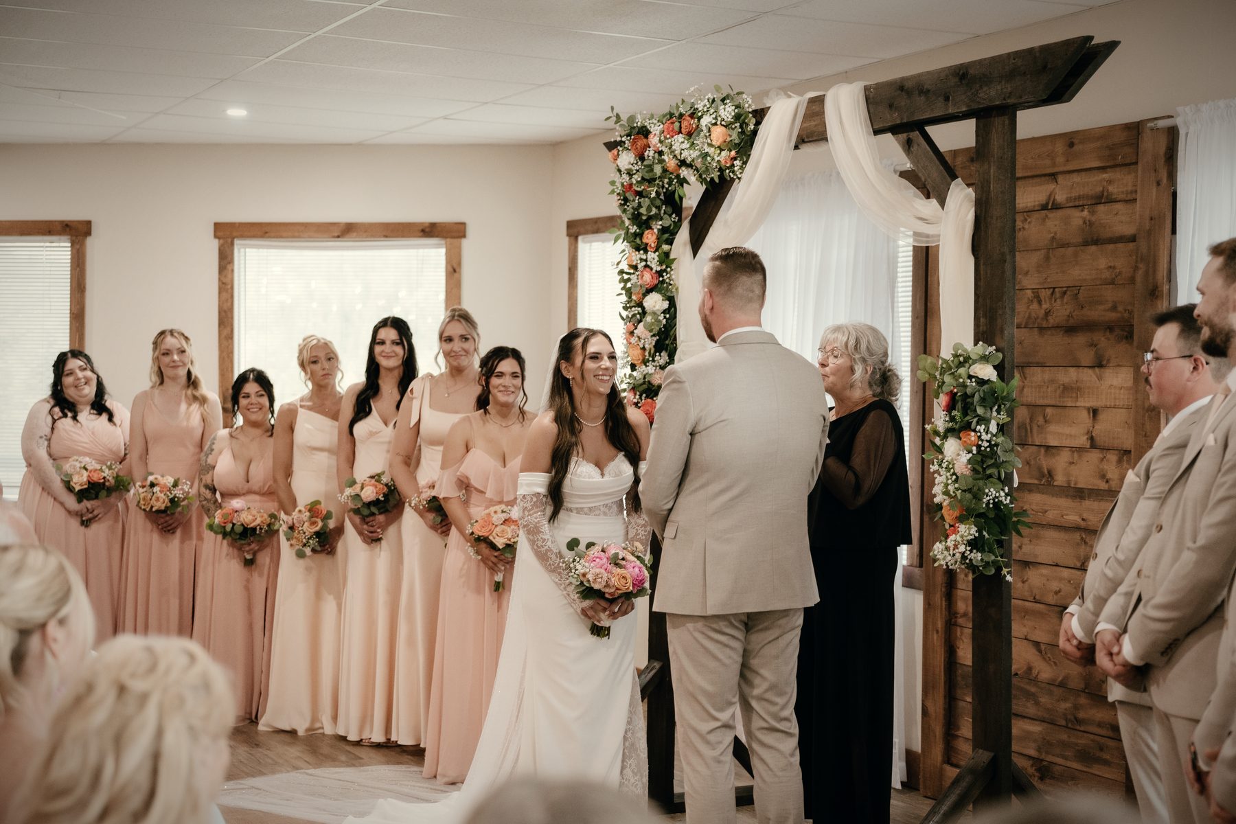The couple and their wedding party at the timber arch in the hall, white drapery behind them and string lights overhead
