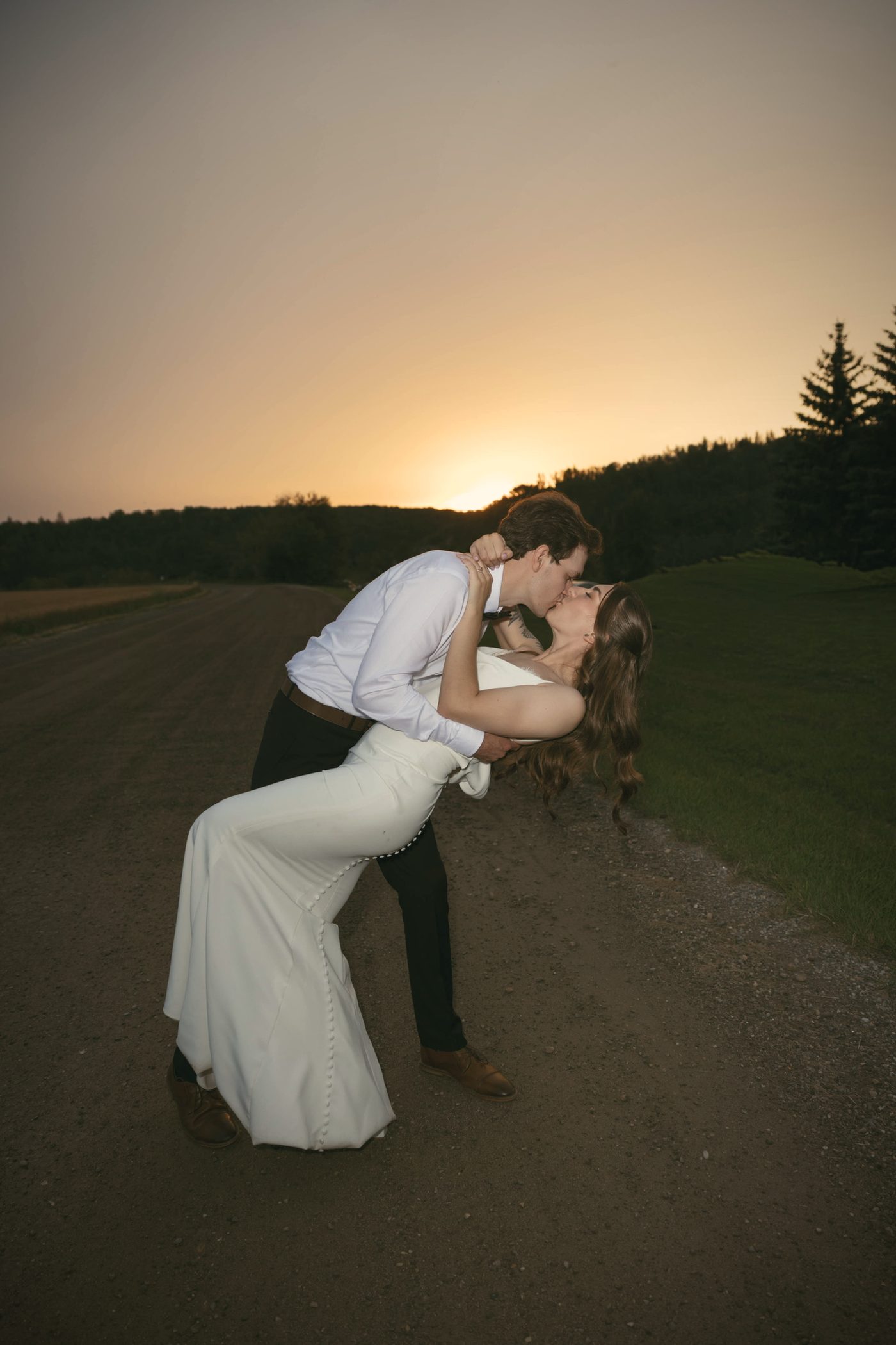 A couple dipping into a kiss on the road at sunset, the sky orange over the hill