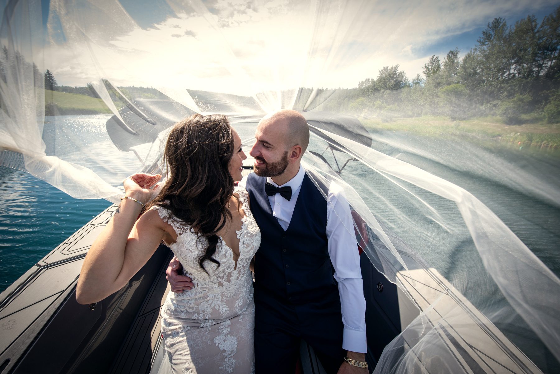 A couple pressed forehead to forehead in the boat, her veil lifting around them over green water
