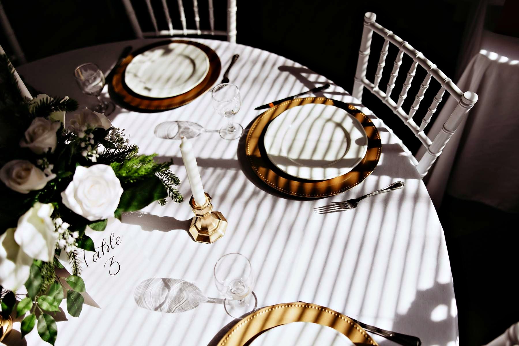 A guest table from above, gold chargers, white roses, low afternoon light across the linen