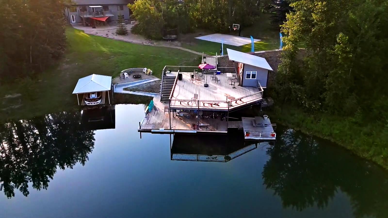 The dock and deck seen from directly above, reflected whole in still water, the family home up the bank behind them