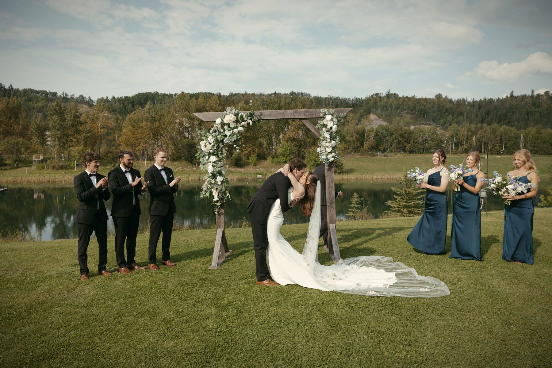 A kiss under the flowered arch, the wedding party and bridesmaids in blue looking on
