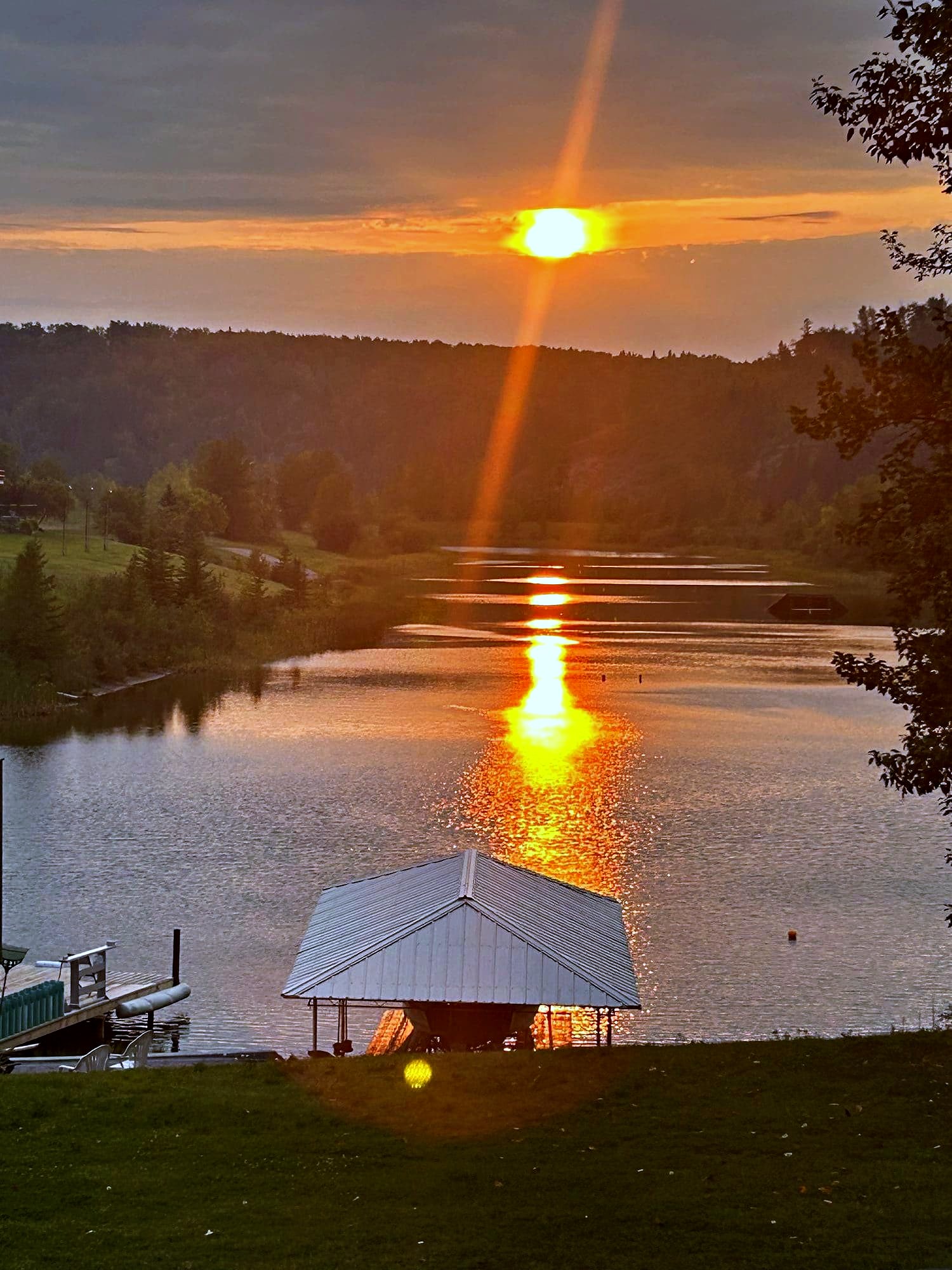 Sunset over the lake, the sun low on the far treeline and its path laid across the water to the boathouse