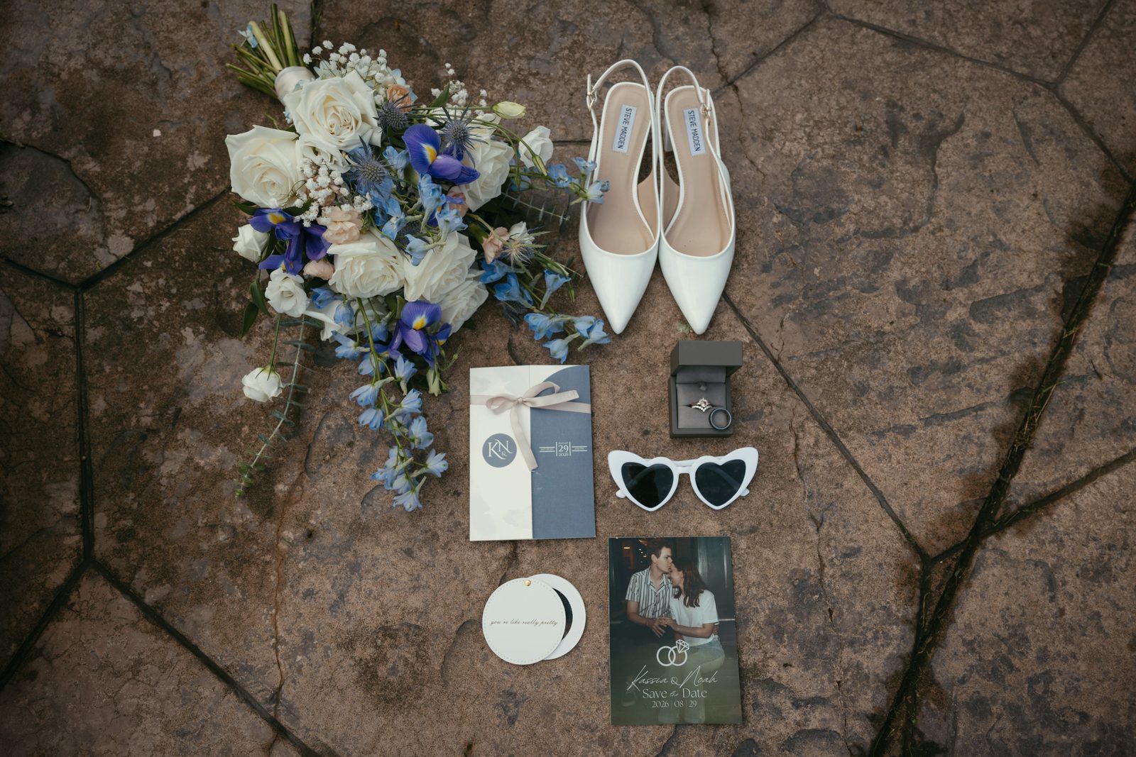 A bouquet, heels, sunglasses, a ring box and stationery laid out on a stone floor