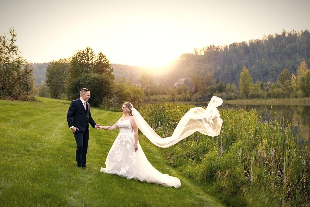 A groom leading his bride across the lawn by the water, her long veil lifting behind them