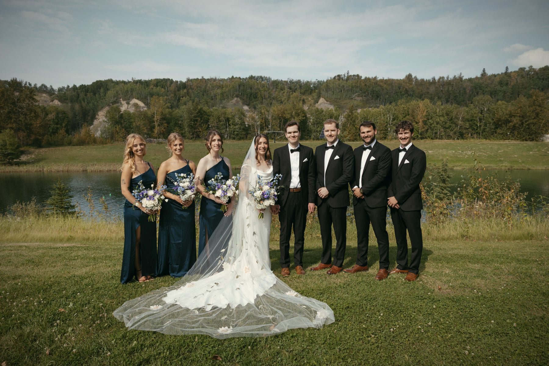 A bride in a long veil with the groom and the wedding party standing on the lawn, the lake behind them