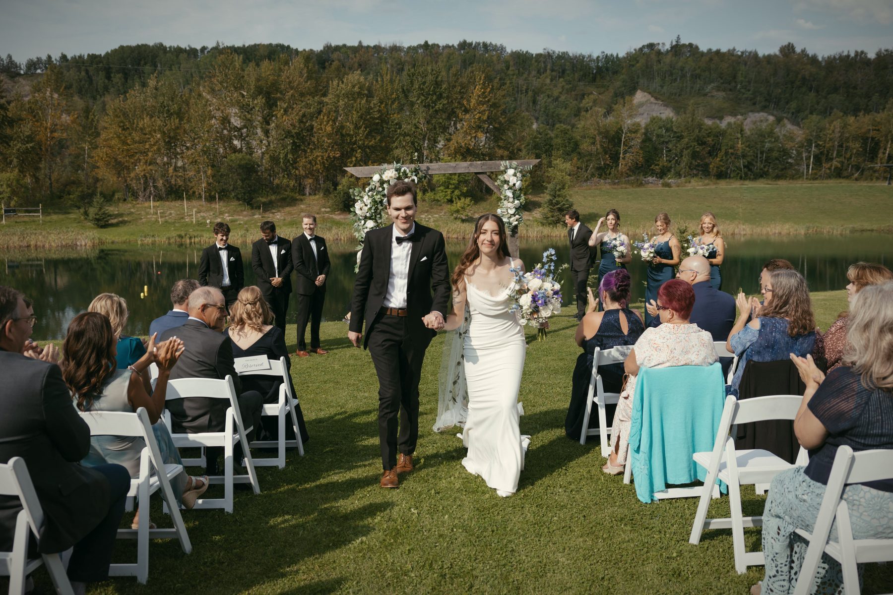 A couple walking back up the aisle between seated guests, the arbour and the lake behind them